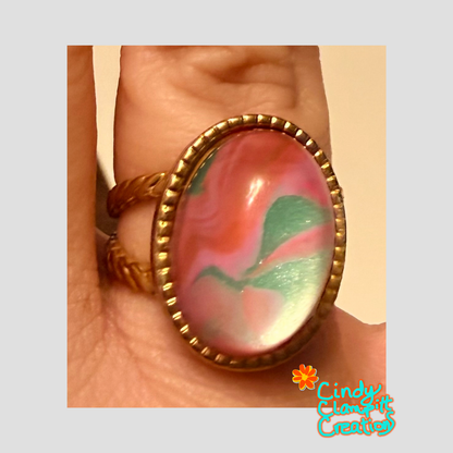Hand-painted pink and green swirl oval glass cabochon ring on a gold twisted rope open cuff band, worn on finger