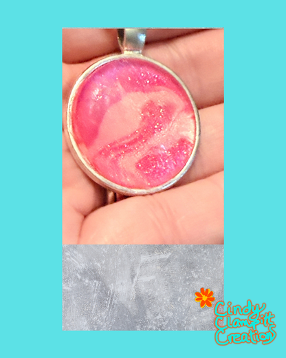 A handmade pink and white glitter pendant in a silver bezel setting, held in a person's hand, with a textured gray overlay at the bottom and a 'Cindy's Creative' logo watermark.