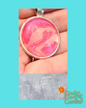 A handmade pink and white glitter pendant in a silver bezel setting, held in a person's hand, with a textured gray overlay at the bottom and a 'Cindy's Creative' logo watermark.