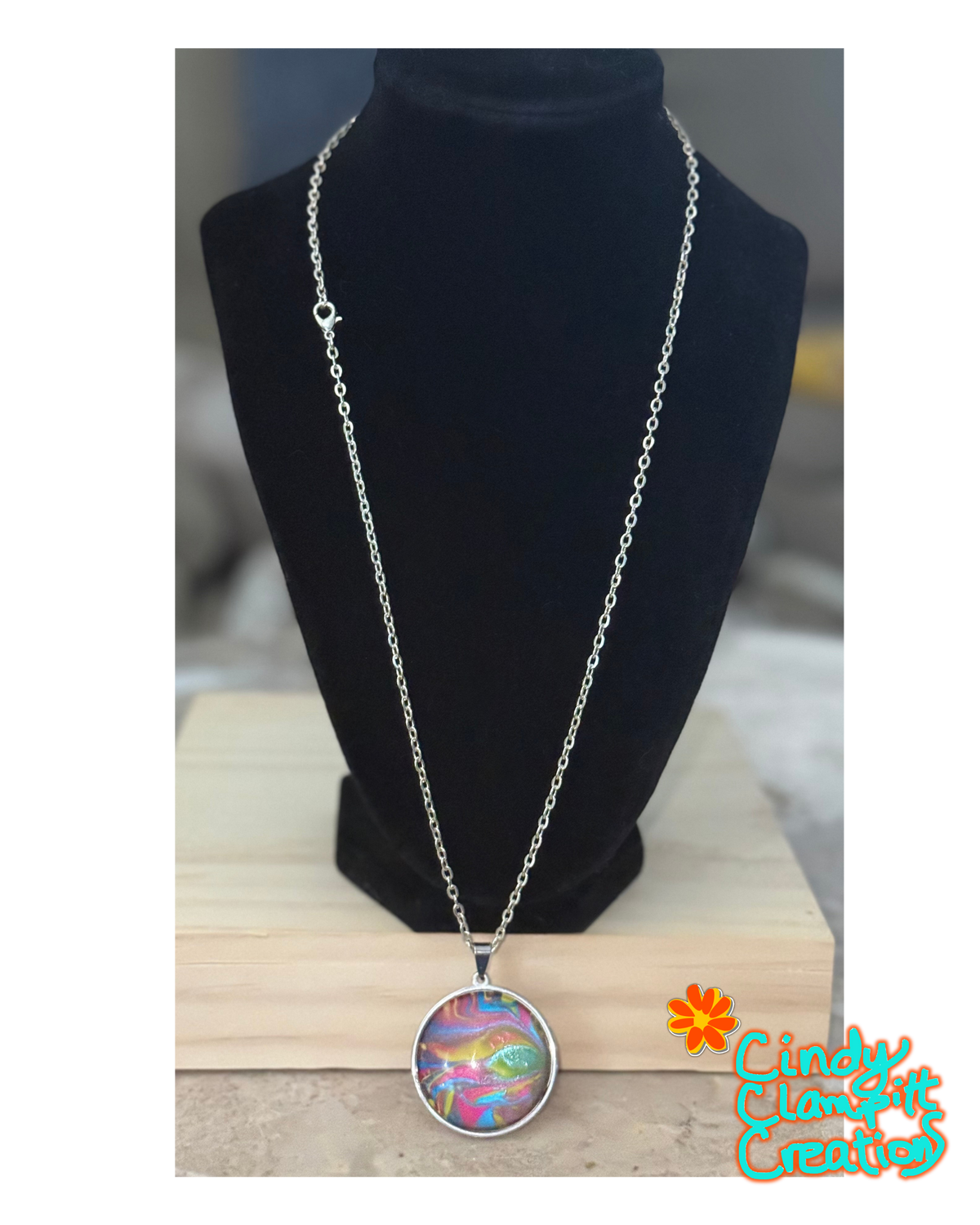 A silver chain necklace with a round pendant featuring an iridescent, multicolored abstract pattern, displayed on a black velvet jewelry bust. The necklace is resting on a light-colored wooden block, with a 'Cindy's Custom Creations' logo in the bottom right corner.