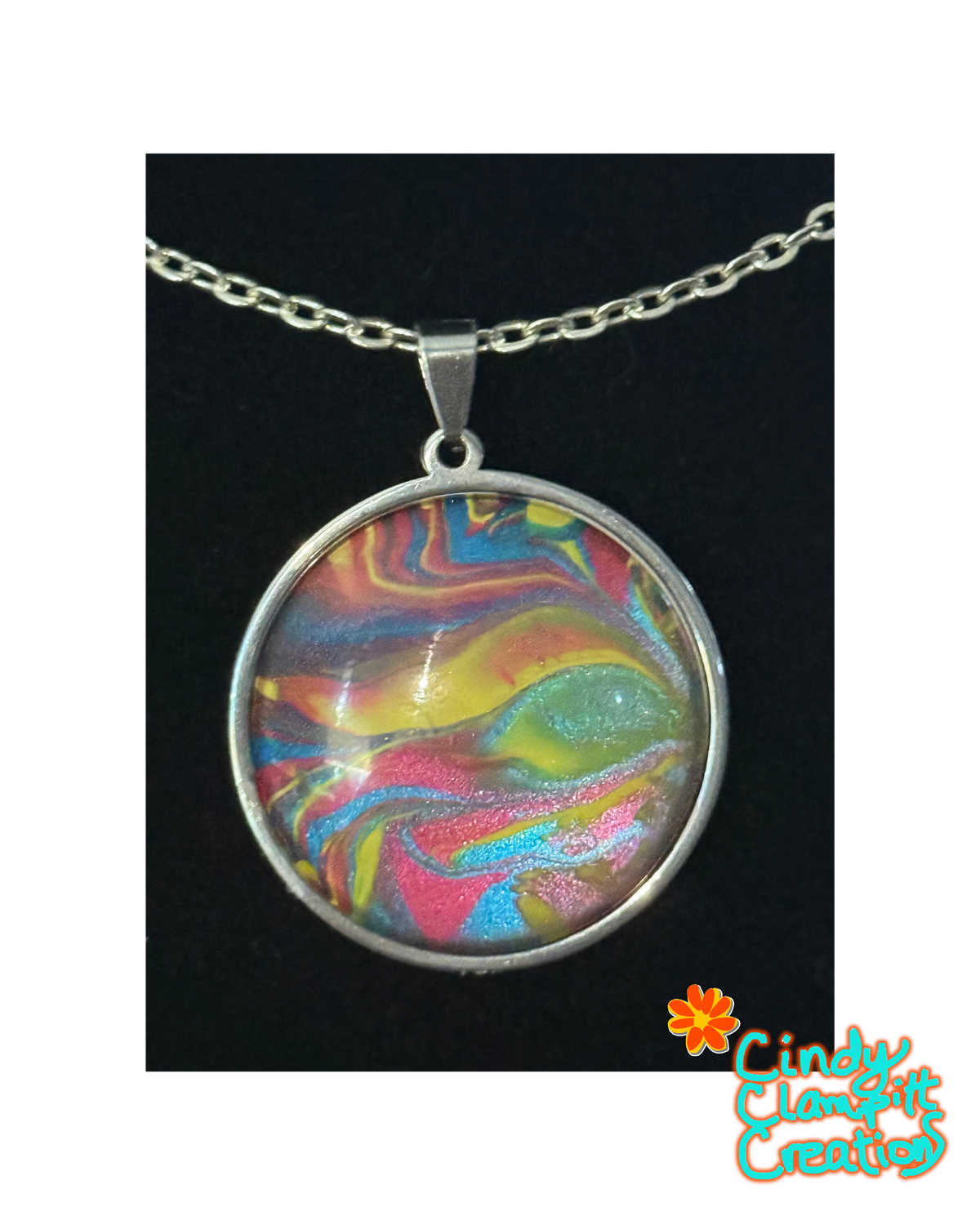 A round silver pendant necklace featuring a colorful marbled swirl design with pink, yellow, blue, and green tones under a clear dome, shown on a black background with a Cindy Clement Creations logo in the bottom right corner.