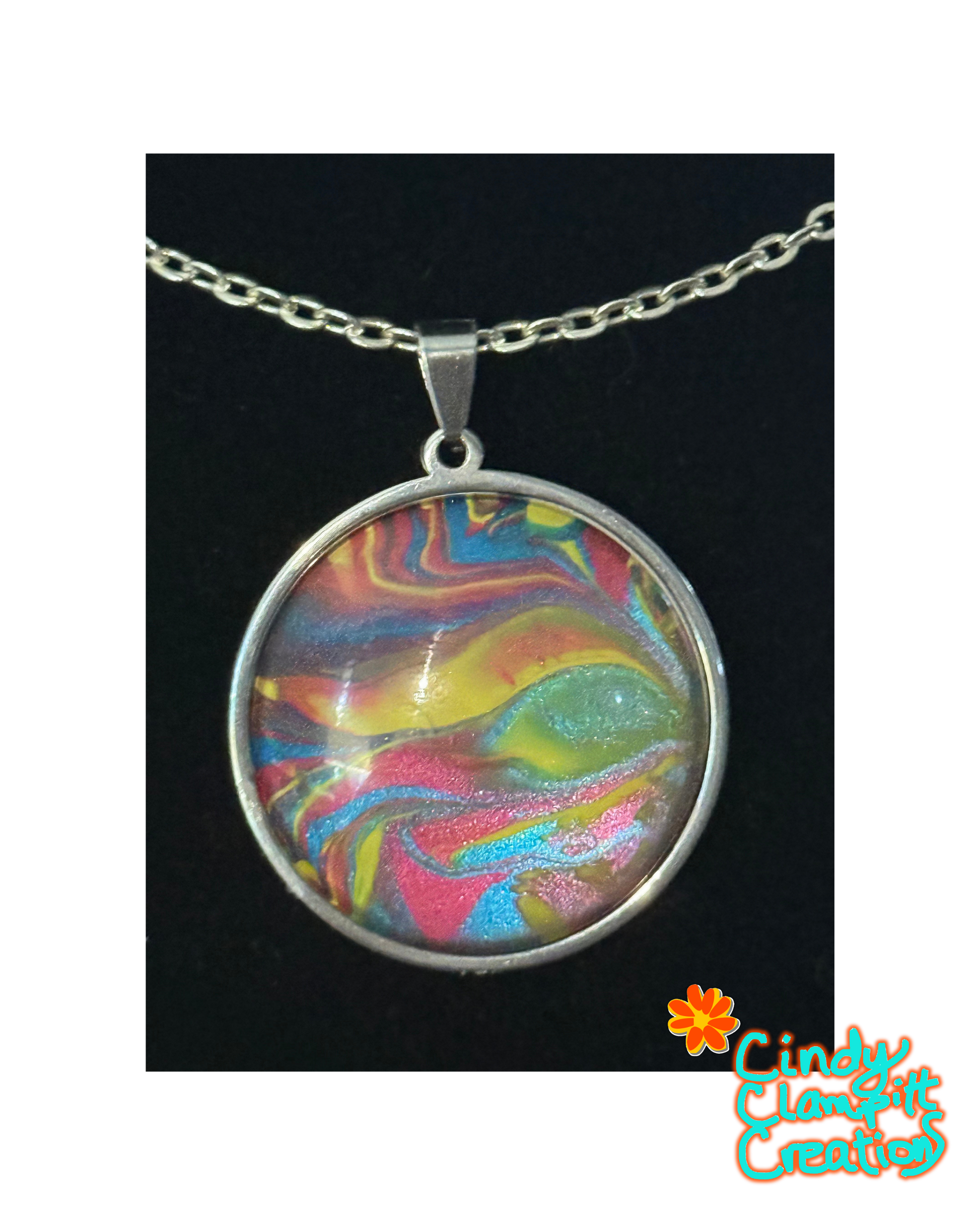 A round silver pendant necklace featuring a colorful marbled swirl design with pink, yellow, blue, and green tones under a clear dome, shown on a black background with a Cindy Clement Creations logo in the bottom right corner.