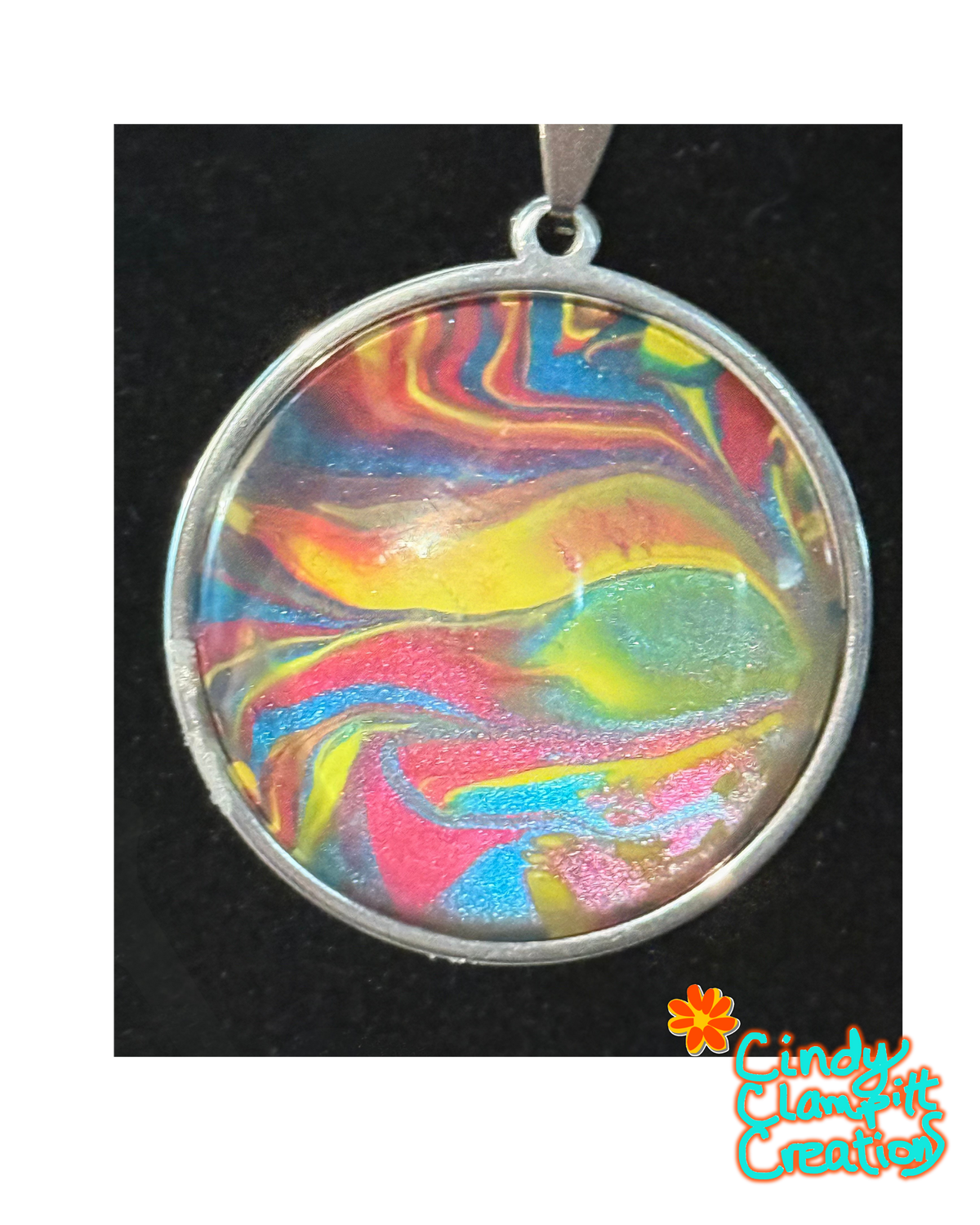 A round silver pendant with a colorful marbled resin design featuring swirls of yellow, pink, blue, and green, set against a black background with a 'Cindy's Classtic Creations' logo in the bottom right corner.